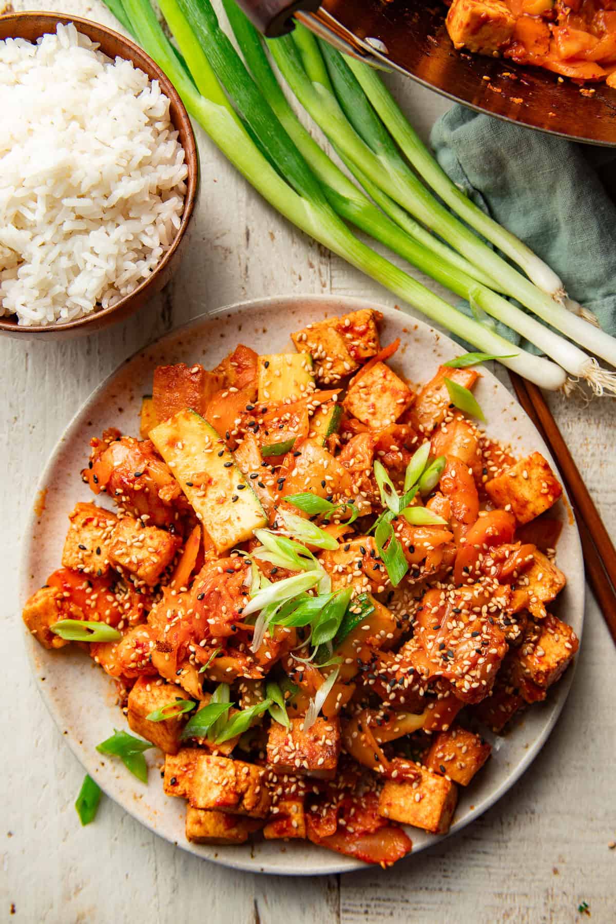 White wooden surface set with plate of Kimchi Stir-Fry, bowl of rice, and bunch of scallions.