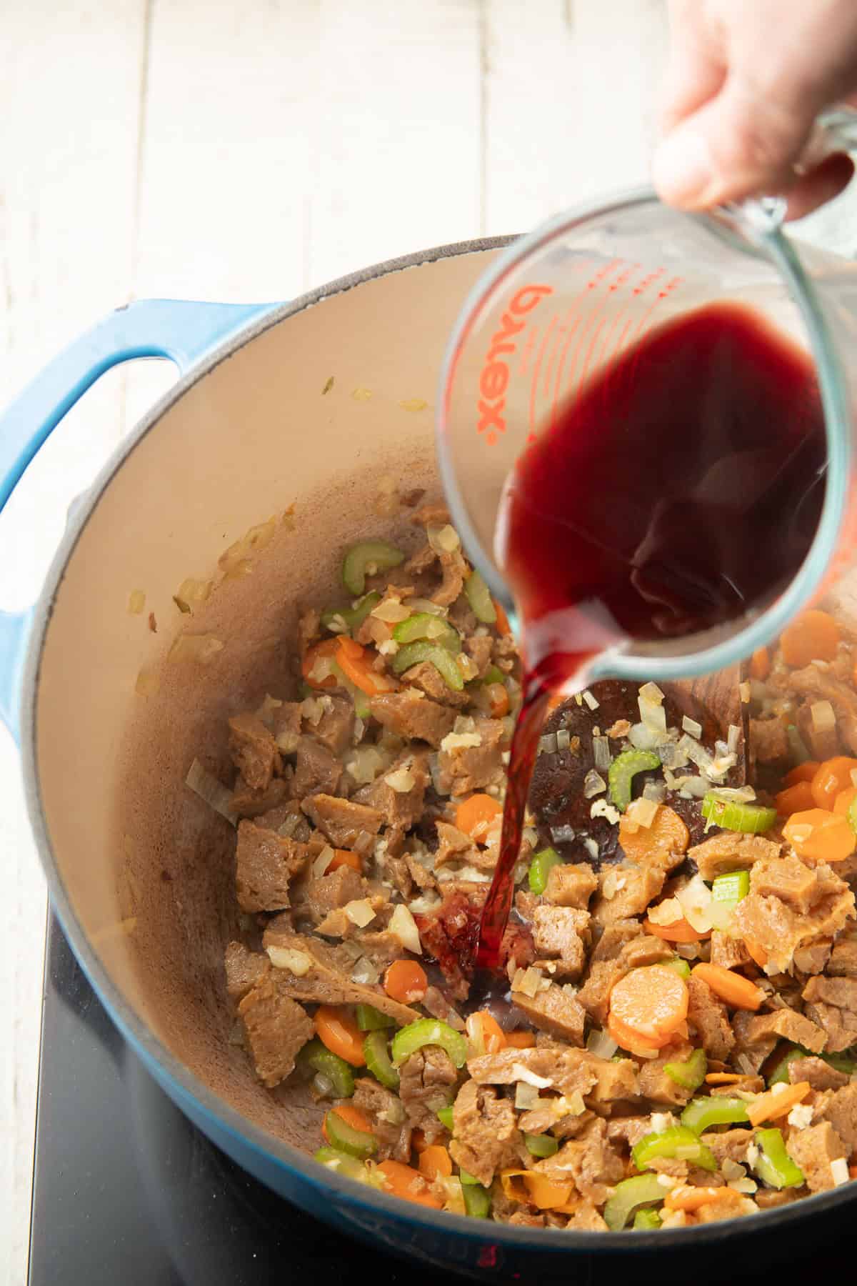Red wine being poured over seitan and vegetables in a pot.