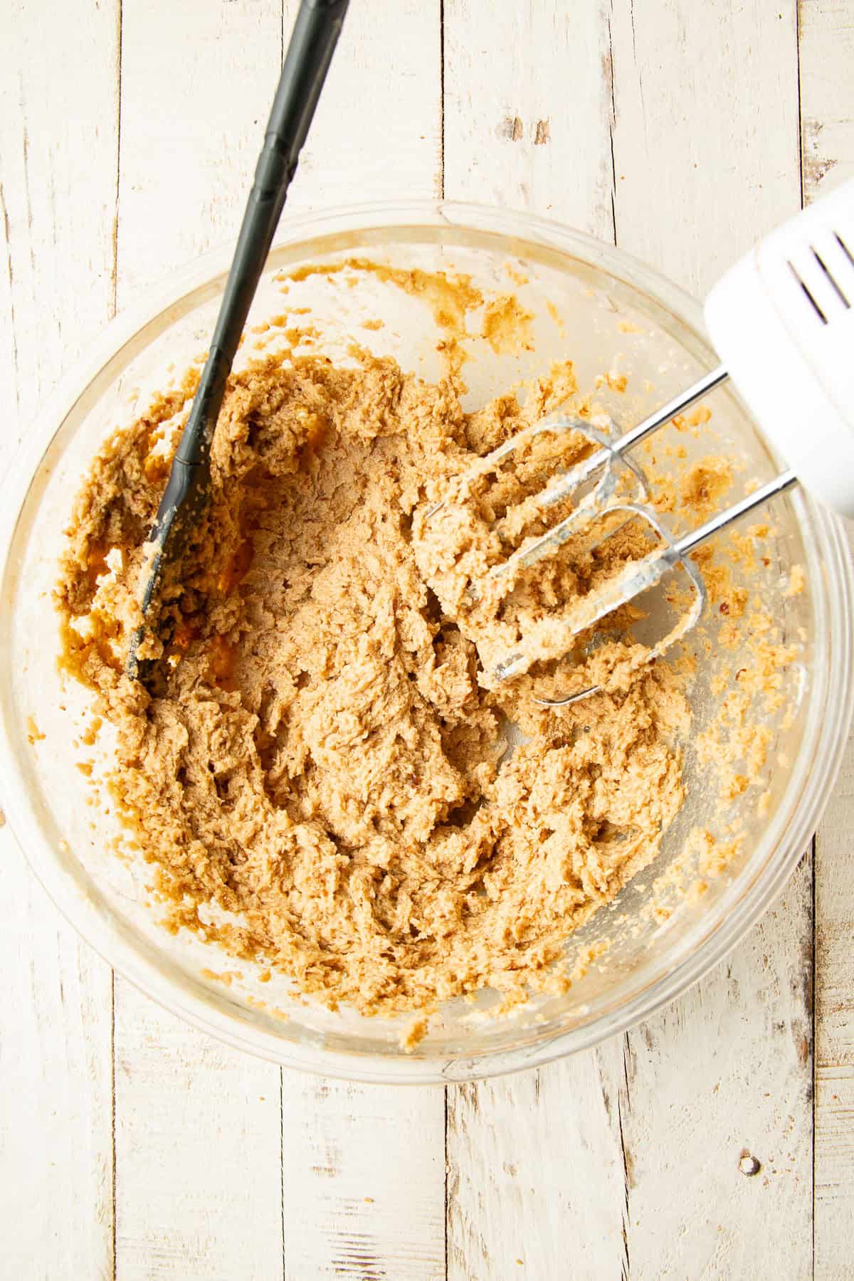 Blend of wet ingredients for cookie dough in a mixing bowl with electric mixer and spatula.
