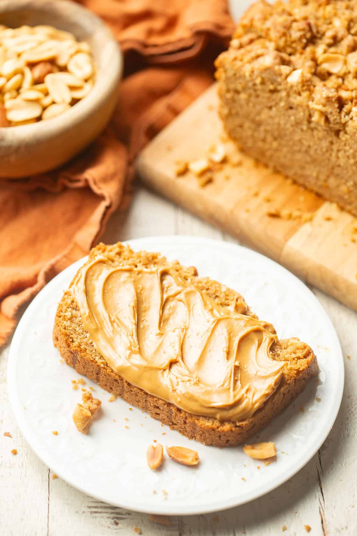 Slice of Peanut Butter Bread with loaf and bowl of peanuts in the background.