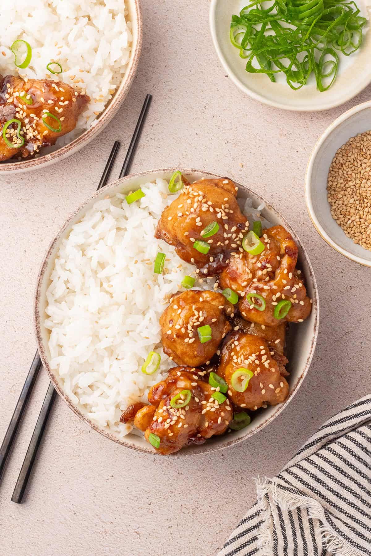 Surface set with two bowls of Sticky Sesame Cauliflower, bowl of scallions, and sesame seeds.