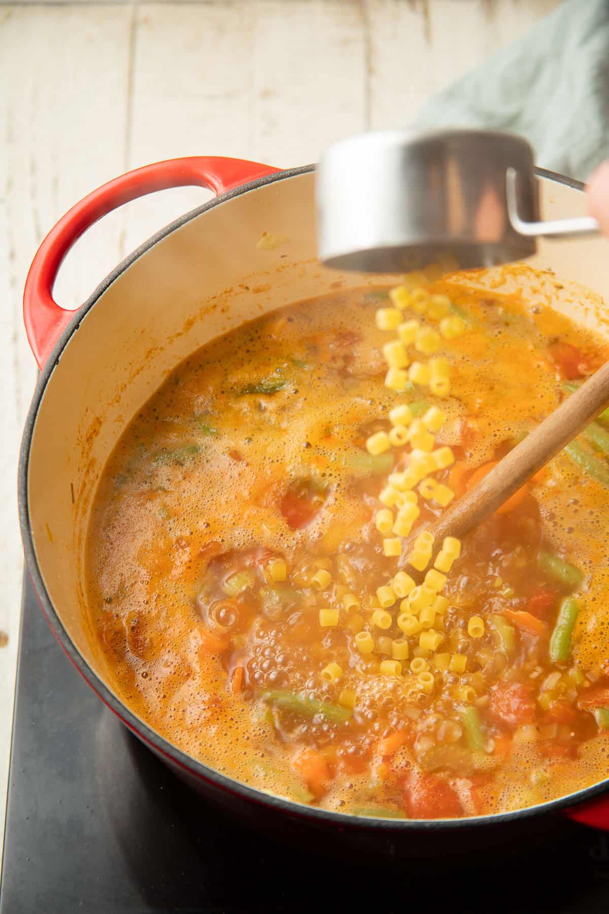 Ditalini pasta being dumped into a pot of simmering vegetable soup.