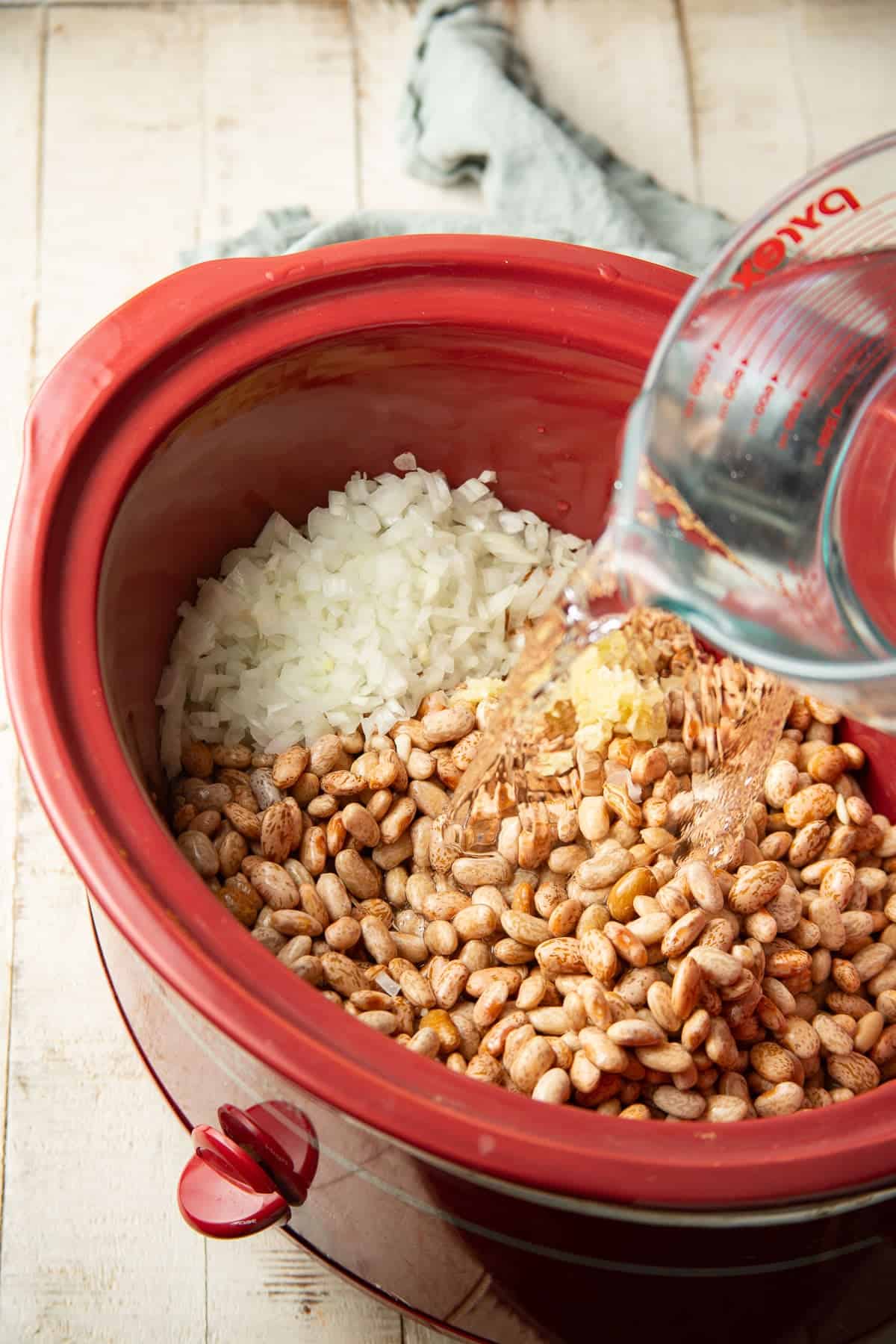 Water being poured over beans and diced onion in a slow cooker.