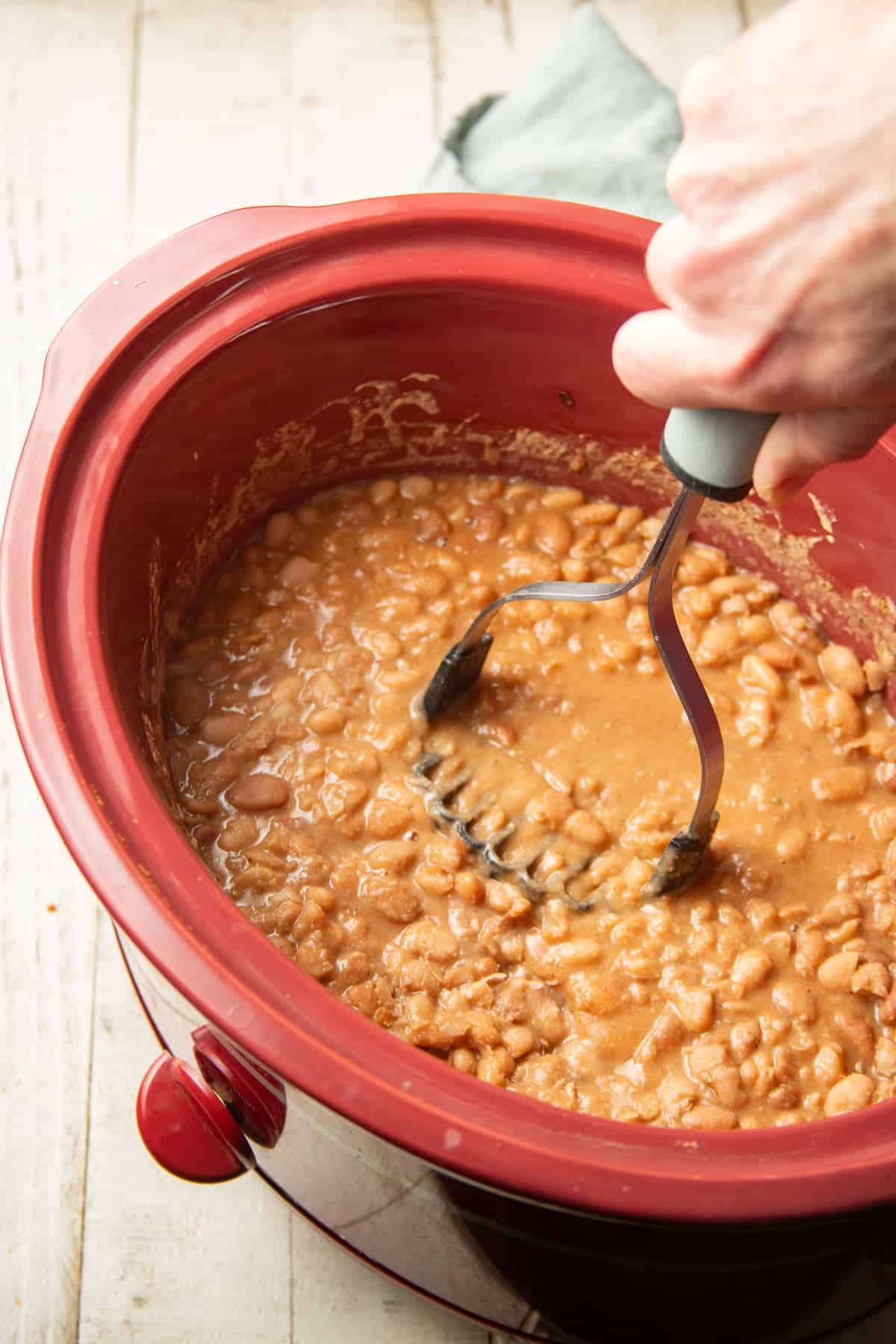 Hand with potato masher mashing cooked beans in a slow cooker.
