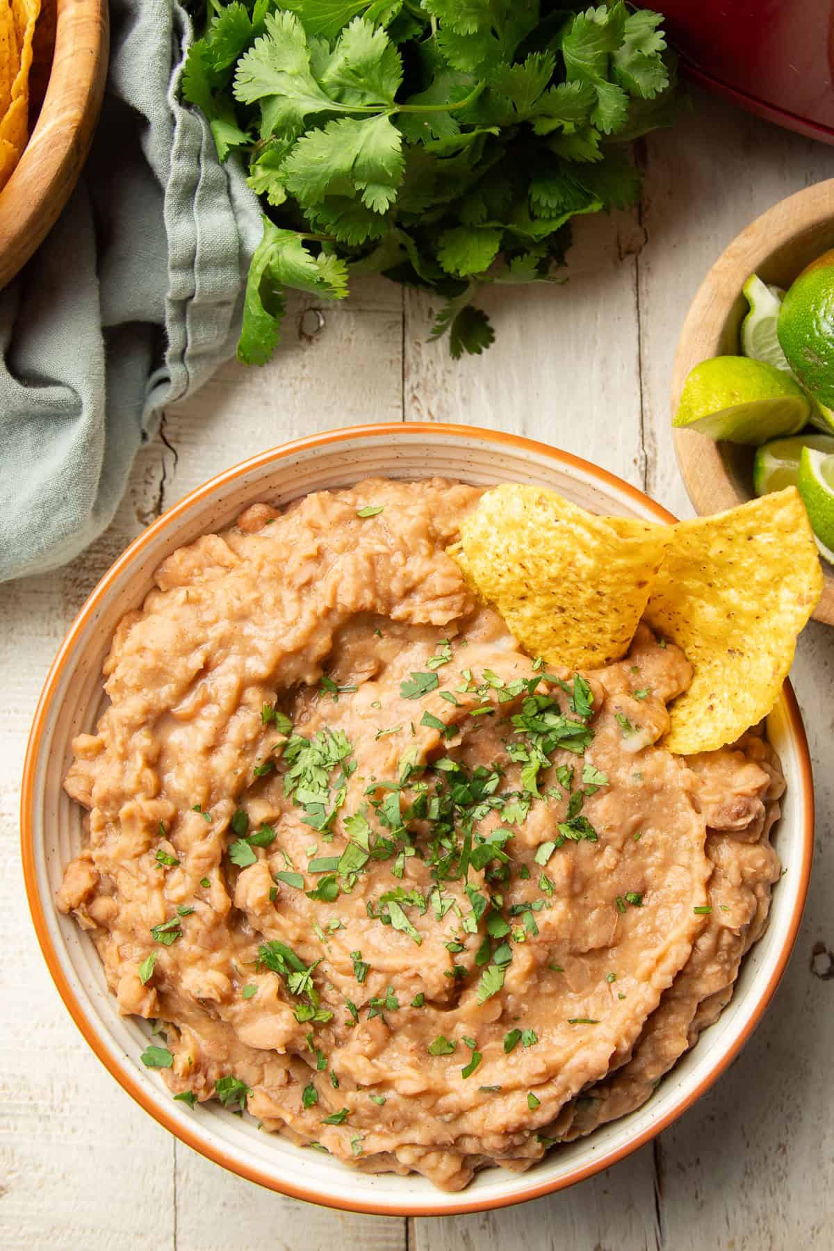 White wooden surface set with a bowl of refried beans, limes, and bunch of cilantro.