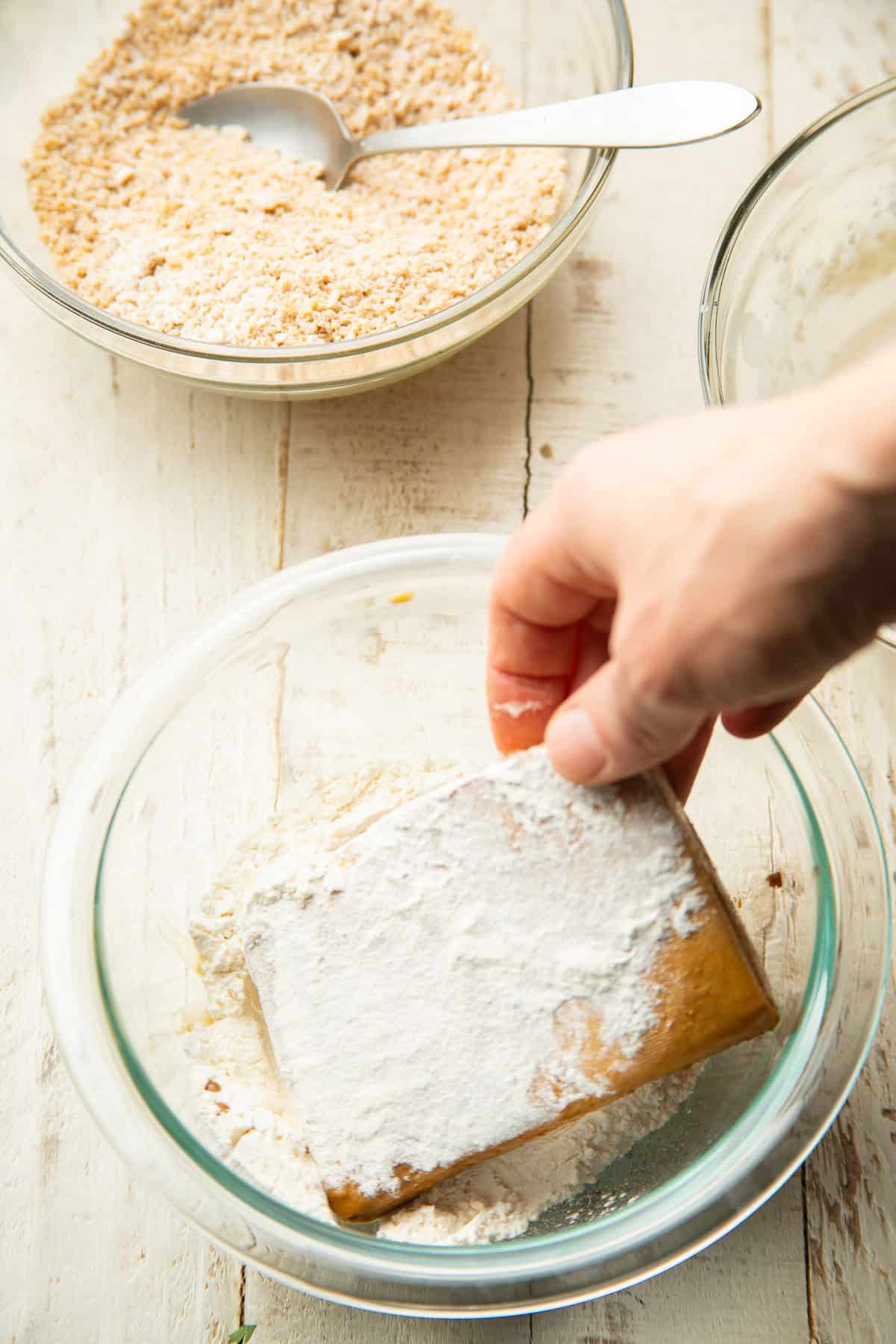 Hand dipping a tofu cutlet in a bowl of flour.