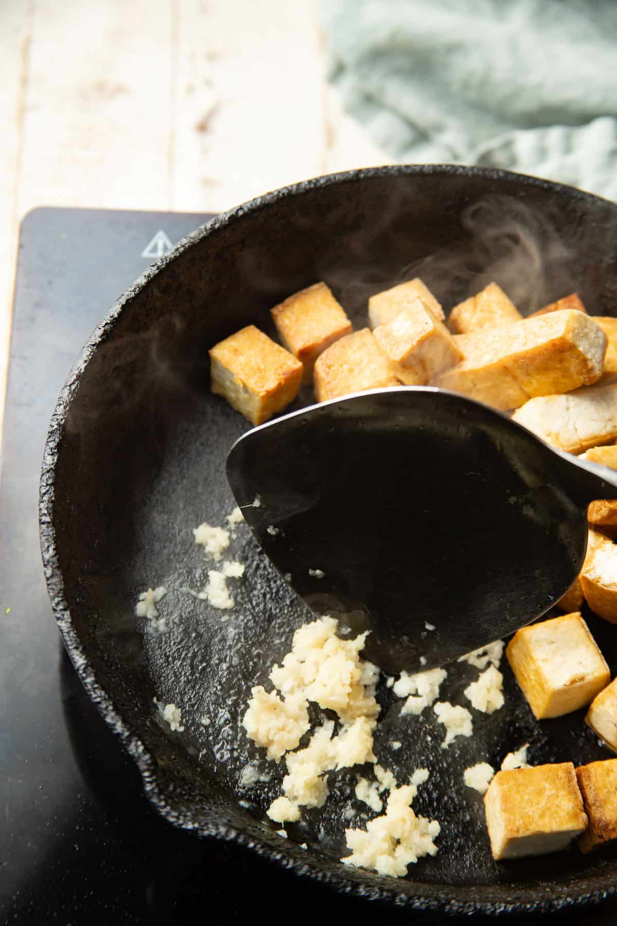 Minced garlic and grated ginger cooking in a skillet with tofu cubes.