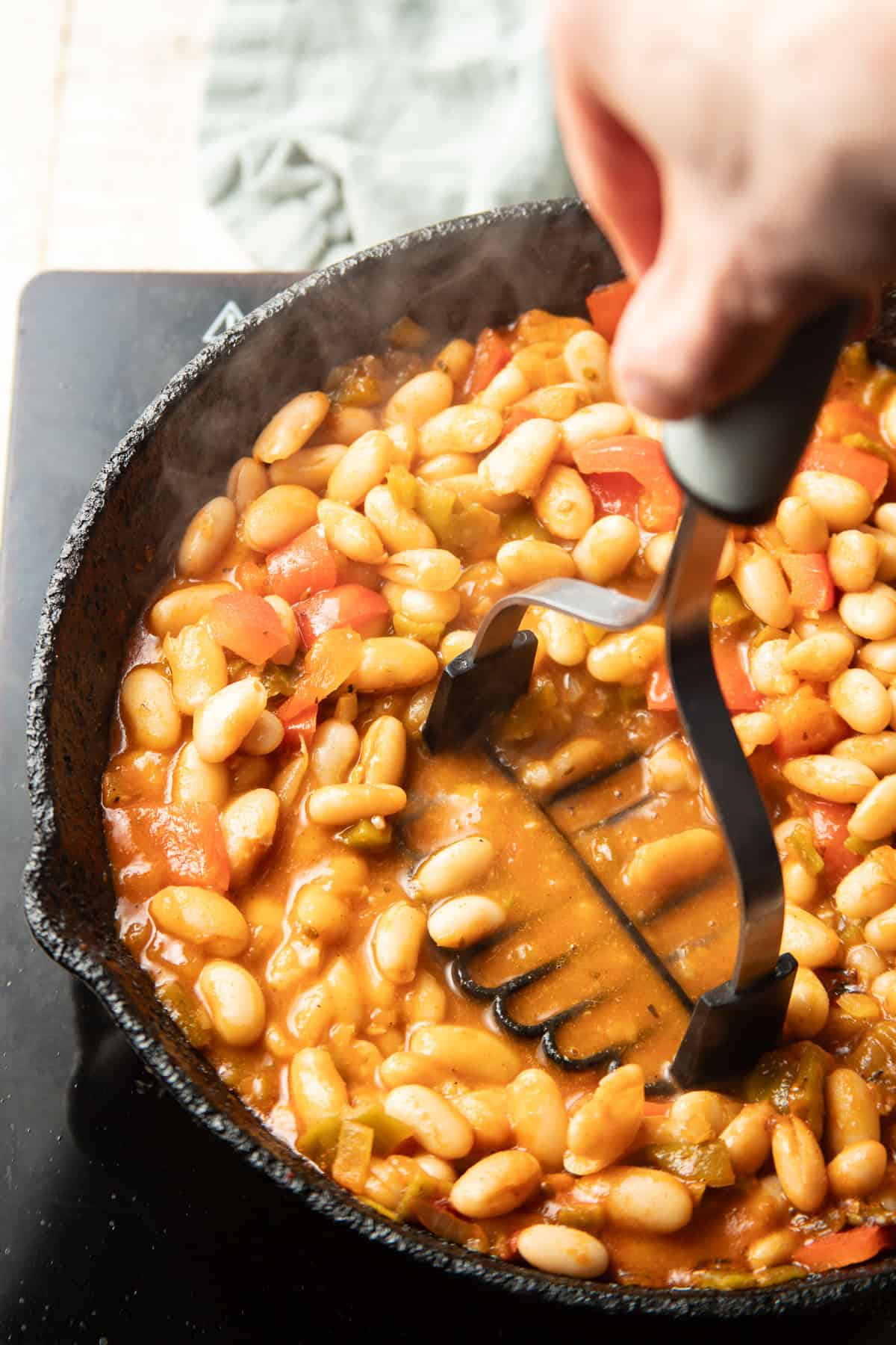Hand with potato masher mashing white beans in a skillet.