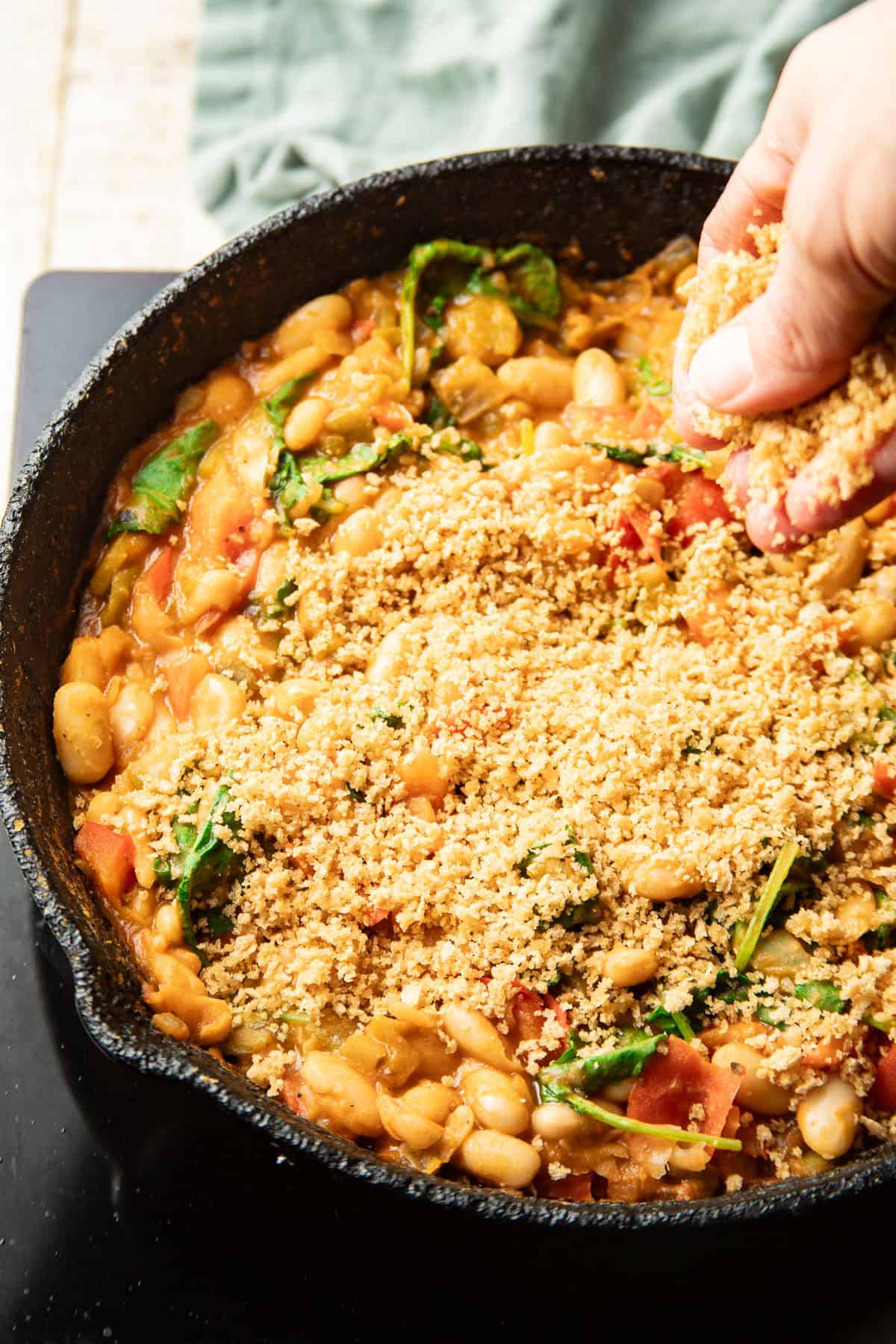 Hand sprinkling panko topping over a White Bean Skillet.