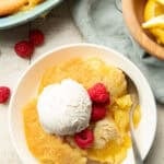White surface set with lemon cobbler, bowl of cobbler, and bowl of lemons.