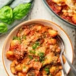 White wooden surface set with baking dish and bowl of Vegan Gnocchi Bake.
