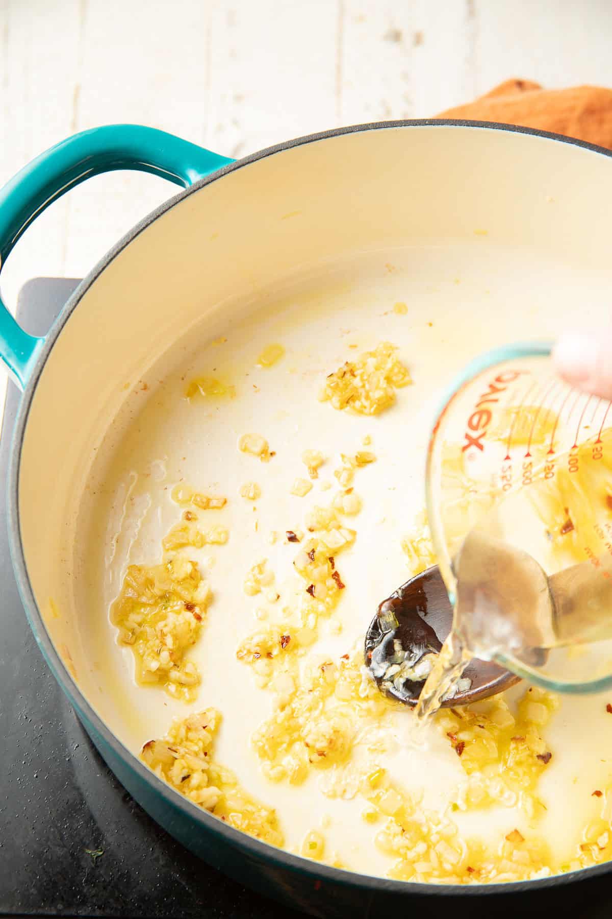 White wine being poured over minced garlic, shallots, and spices in a shallow pot.