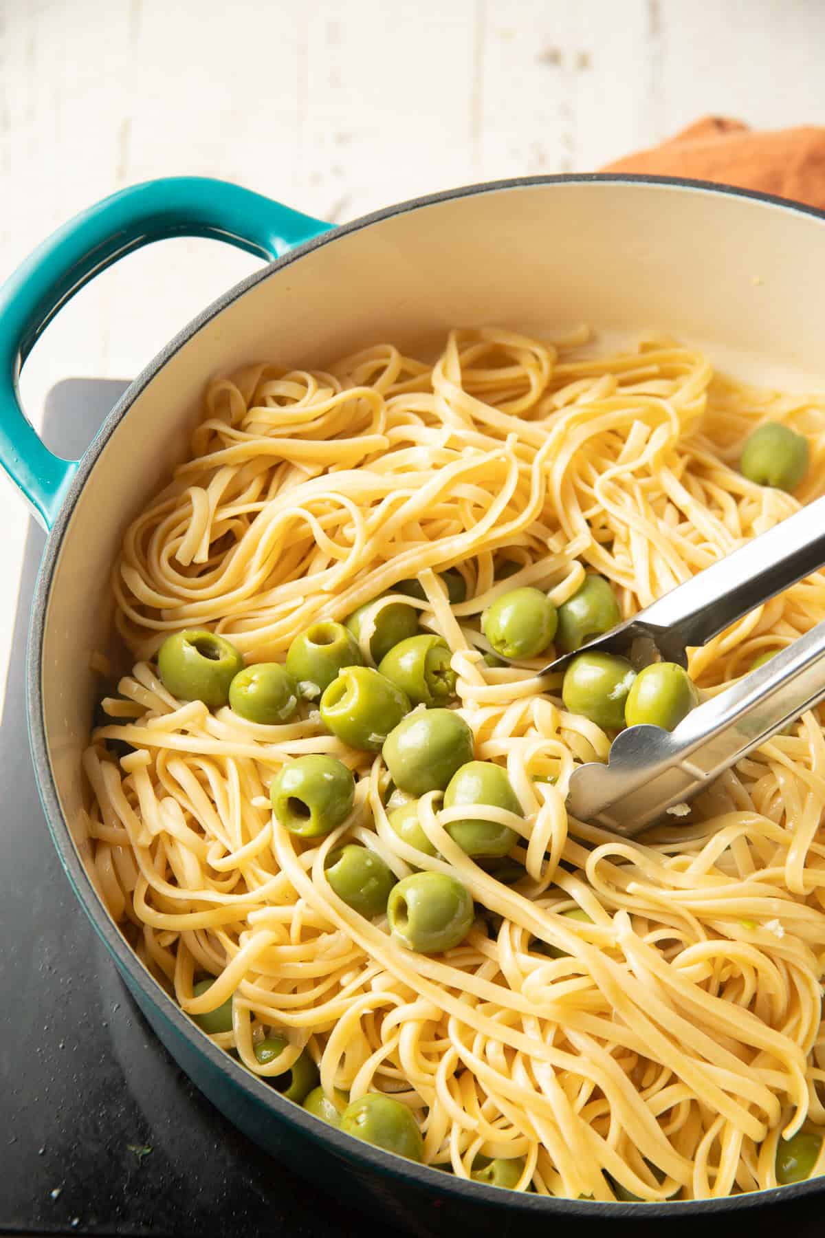 Tongs tossing olives and pasta in a pot.
