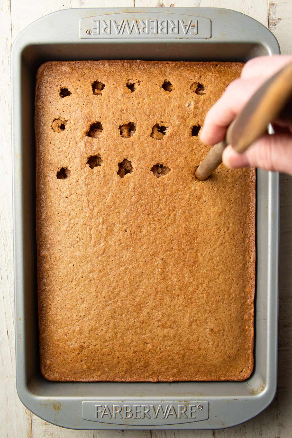 Hand with wooden spoon poking holes in a cake.