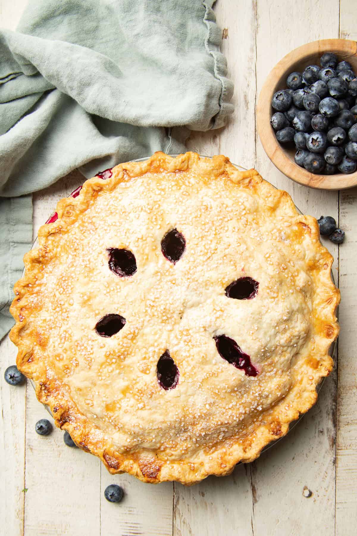 White wooden surface set with a Vegan Blueberry Pie and bowl of blueberries.