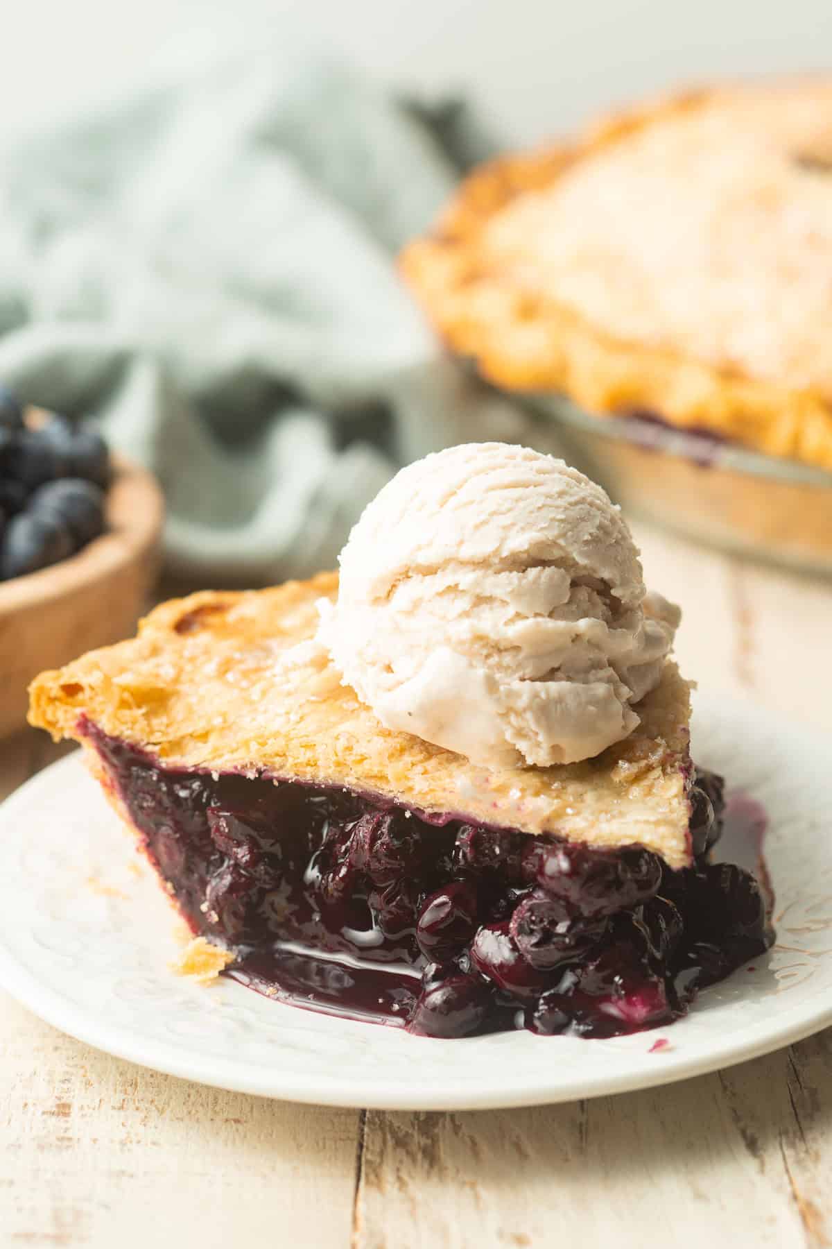 Slice of Vegan Blueberry Pie with pie and bowl of blueberries in the background.