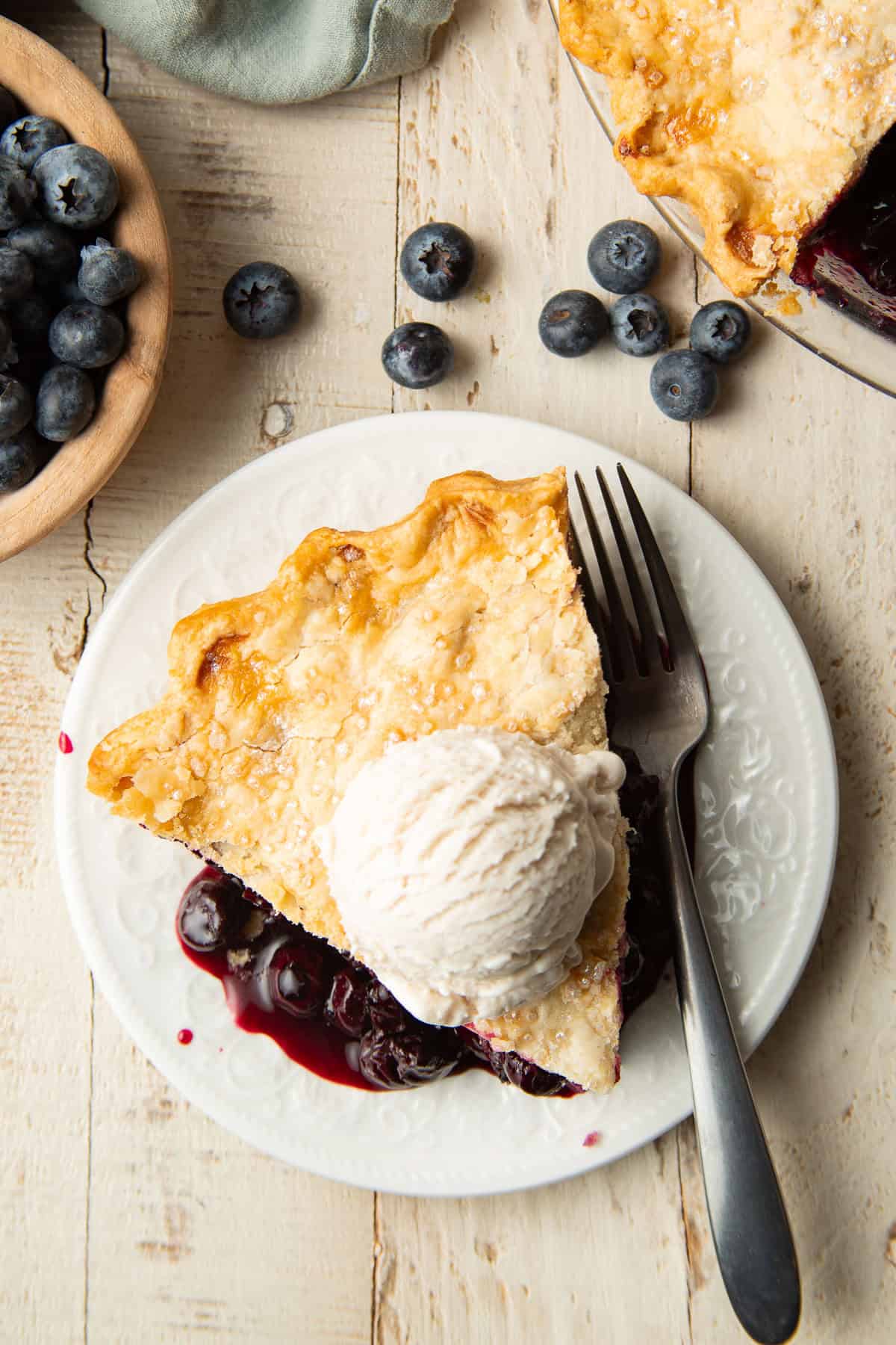 White wooden surface set with Vegan Blueberry Pie, slice of pie, and bowl of blueberries.