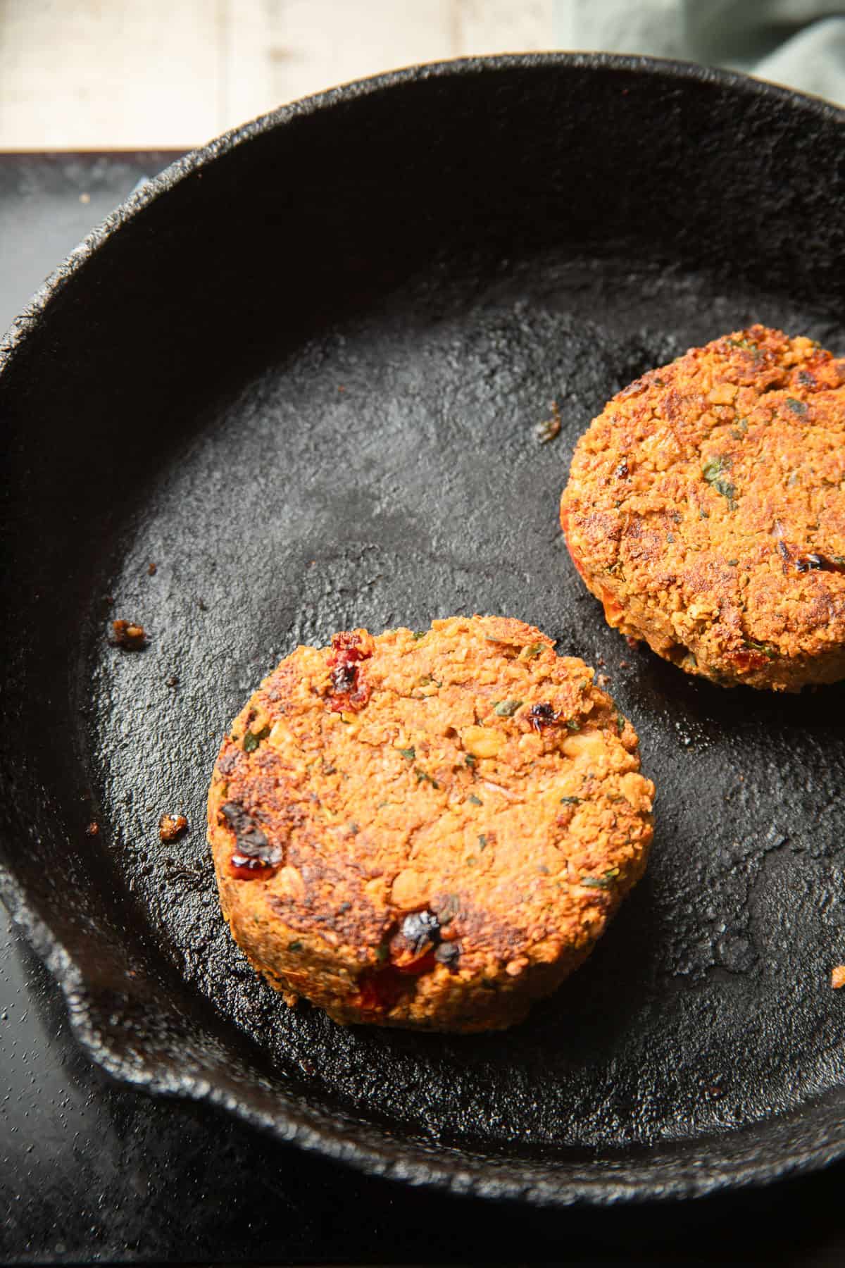 Two Marry Me Chickpea Burger patties cooking in a skillet.