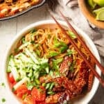 White wooden surface set with skillet, Tempeh Noodle Bowl, and bowl of limes.