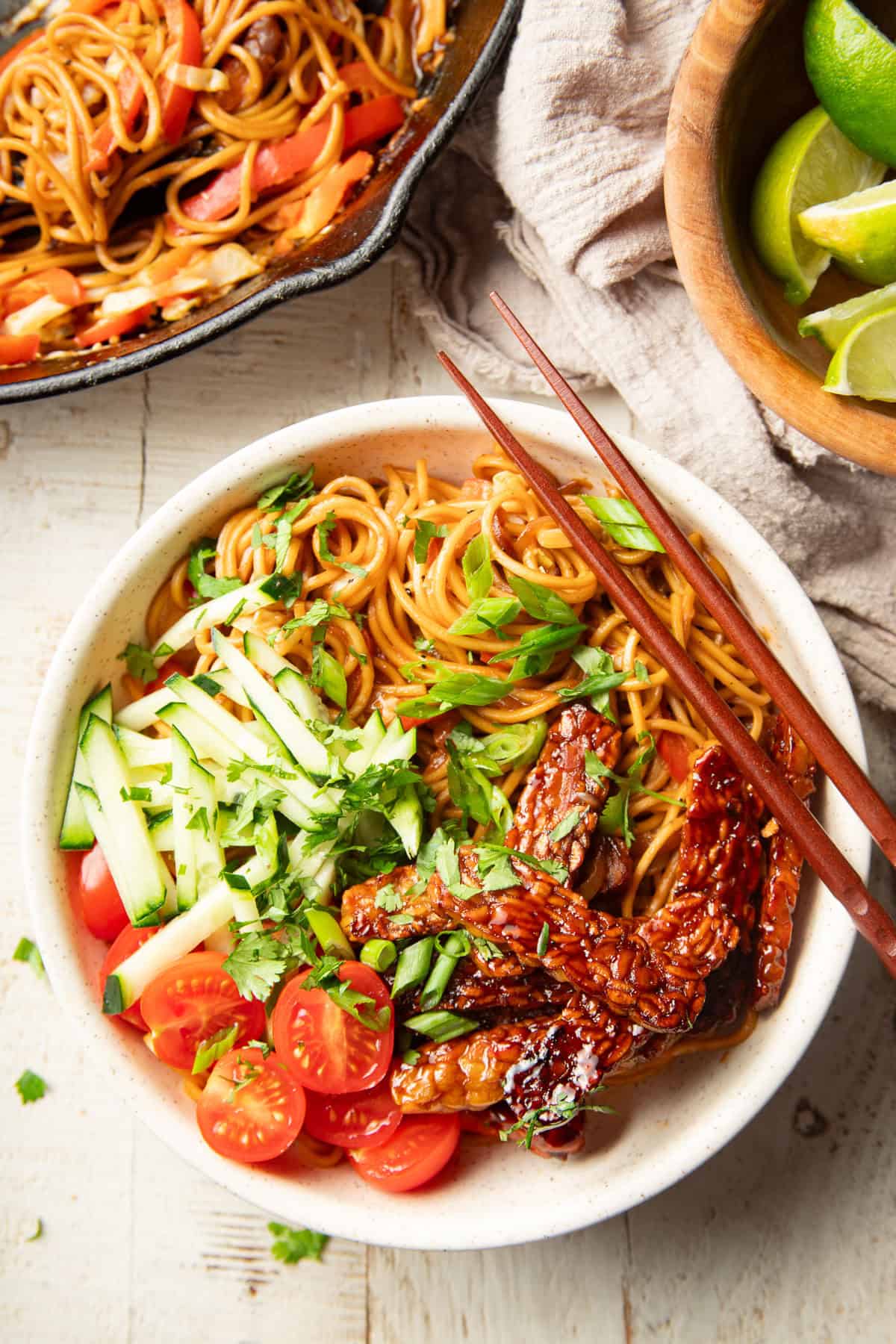 White wooden surface set with skillet, Tempeh Noodle Bowl, and bowl of limes.