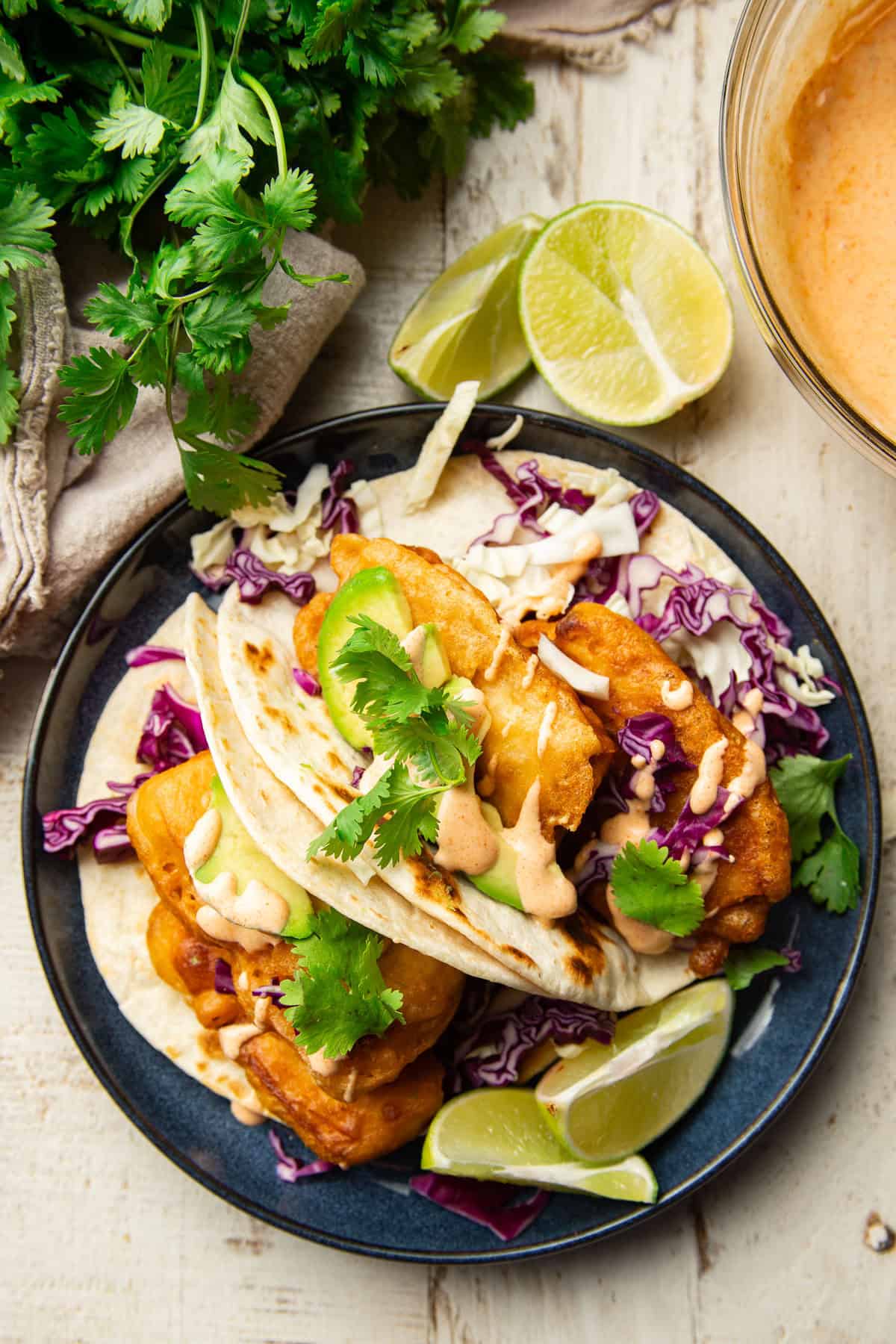 White wooden surface set with plate of Vegan Beer-Battered Fish Tacos, bowl of Baja sauce, and cilantro bunch.