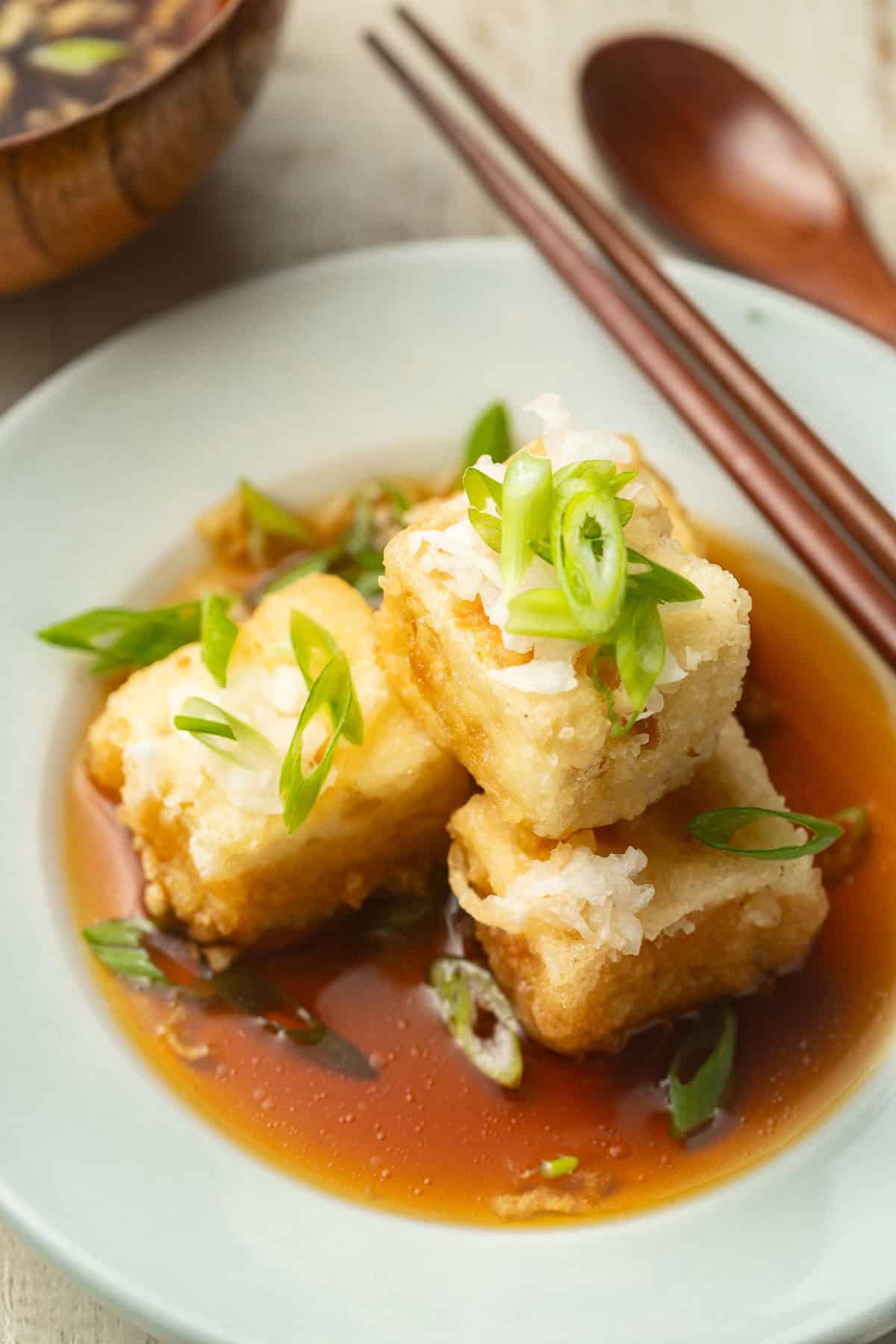 Vegan Agedashi Tofu on a plate with chopsticks, bowl, and spoon in the background.