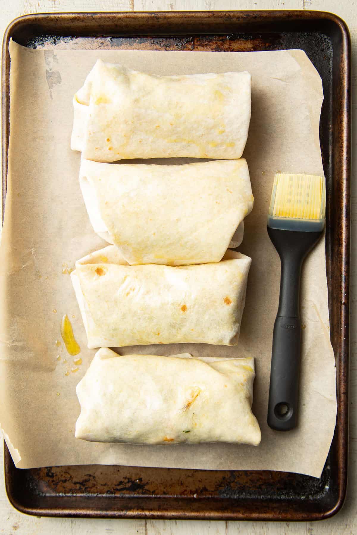 Four unbaked Butternut Squash Black Bean Chimichangas on a baking sheet with basting brush.