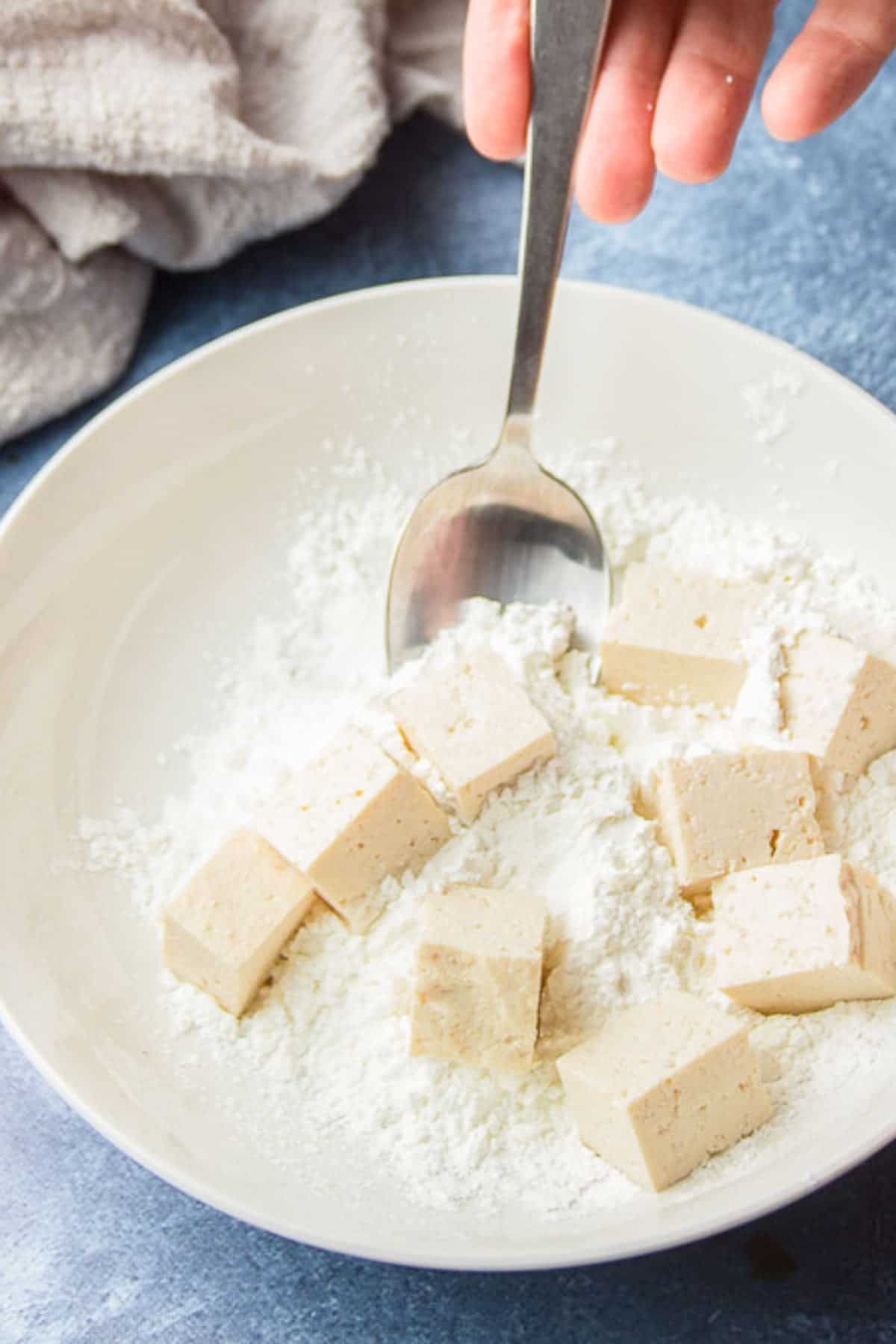 Hand with spoon tossing tofu cubes in a bowl of cornstarch.