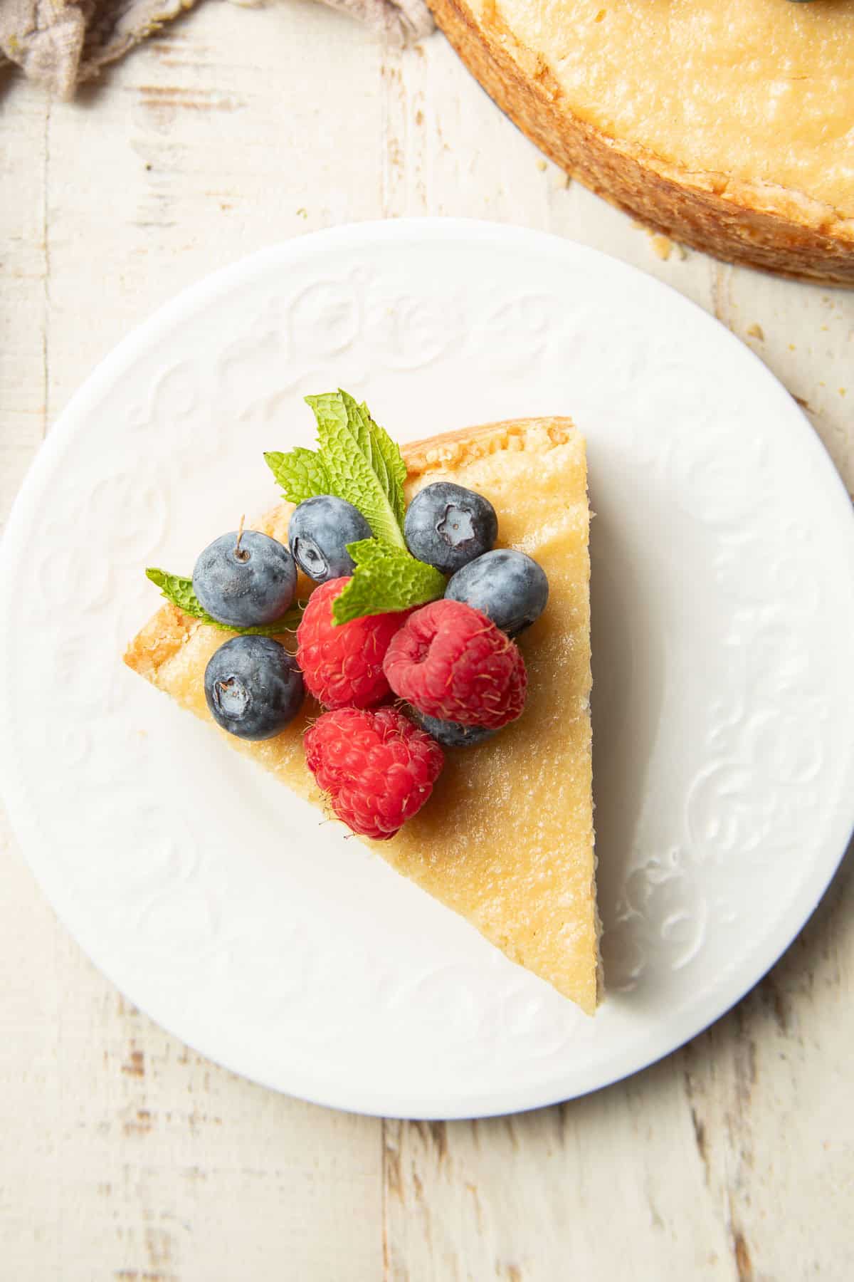 White wooden surface set with slice of Vegan Ricotta Cheesecake on a plate.