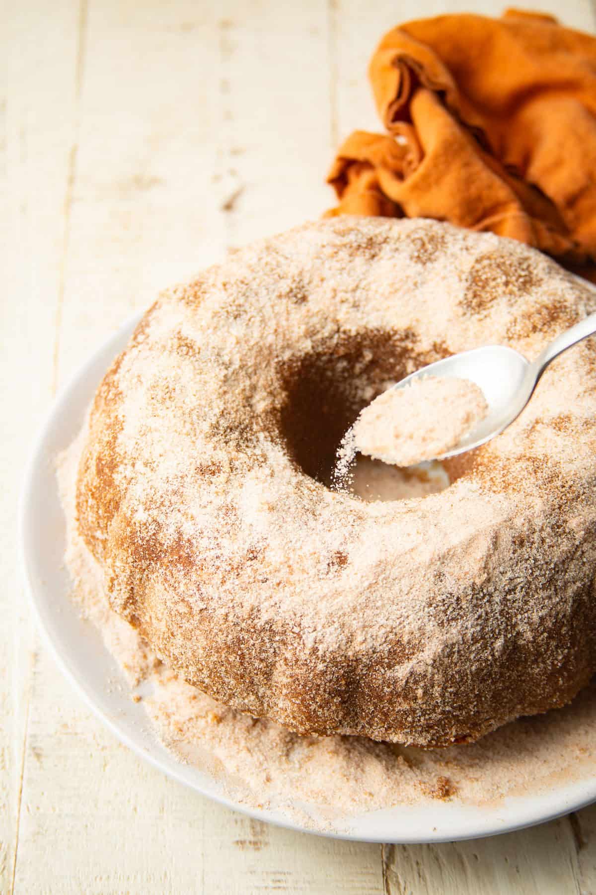Spoon sprinkling cinnamon sugar over an Apple Cider Doughnut Cake.