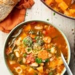 White wooden surface set with a bowl and pot of Fall Minestrone and loaf of bread.