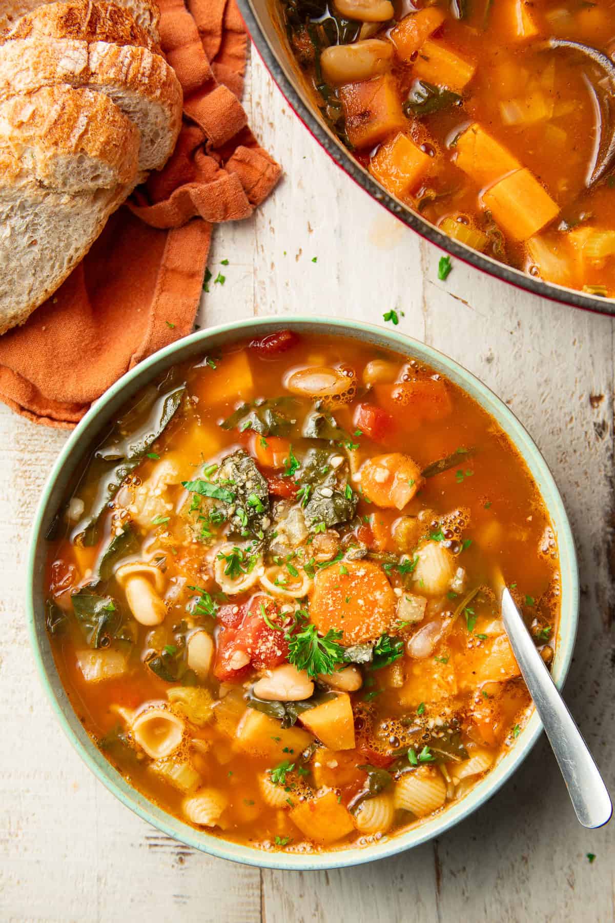 White wooden surface set with a bowl and pot of Fall Minestrone and loaf of bread.