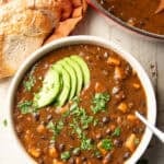 White wooden surface set with pot and bowl of Sweet Potato Black Bean Soup and a loaf of bread.