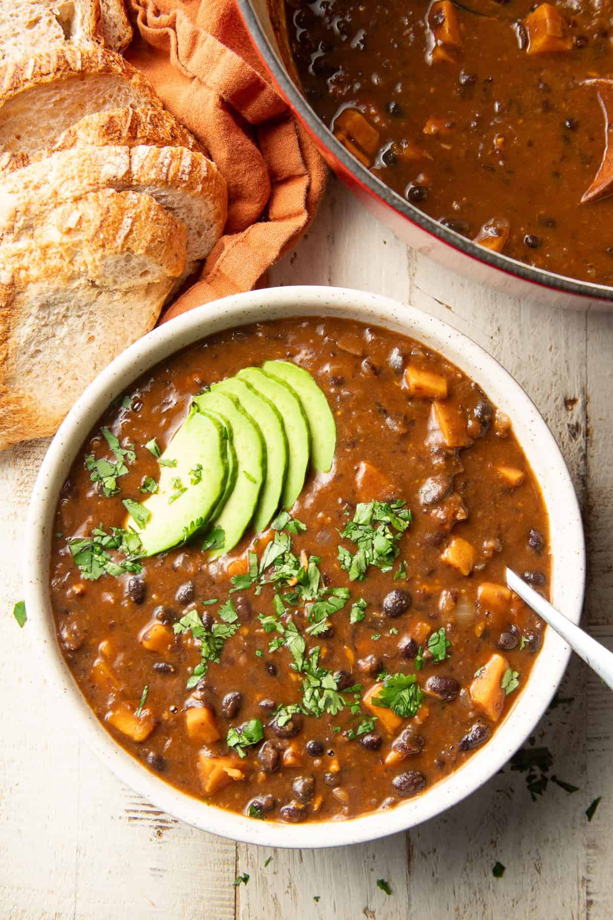 White wooden surface set with pot and bowl of Sweet Potato Black Bean Soup and a loaf of bread.